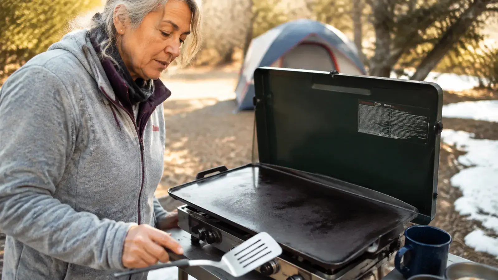 A cast iron griddle on a two-burner camp stove with the center clearly darker and more scorched than the outer edges