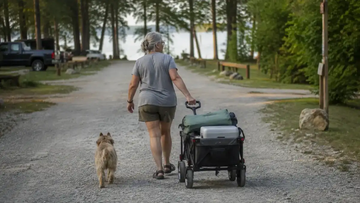 An older woman pulling a loaded collapsible utility wagon down a gravel path toward a walk-in campsite near the water, a mutt dog trotting alongside
