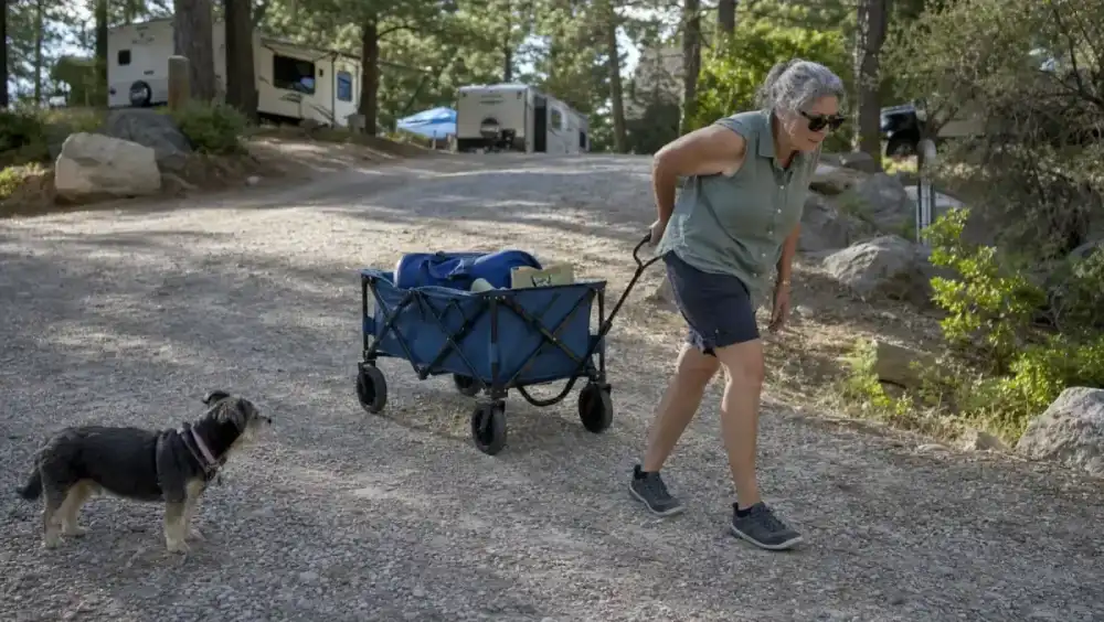 A retired woman straining slightly as she pulls a loaded blue folding utility wagon up a gentle gravel incline toward her truck