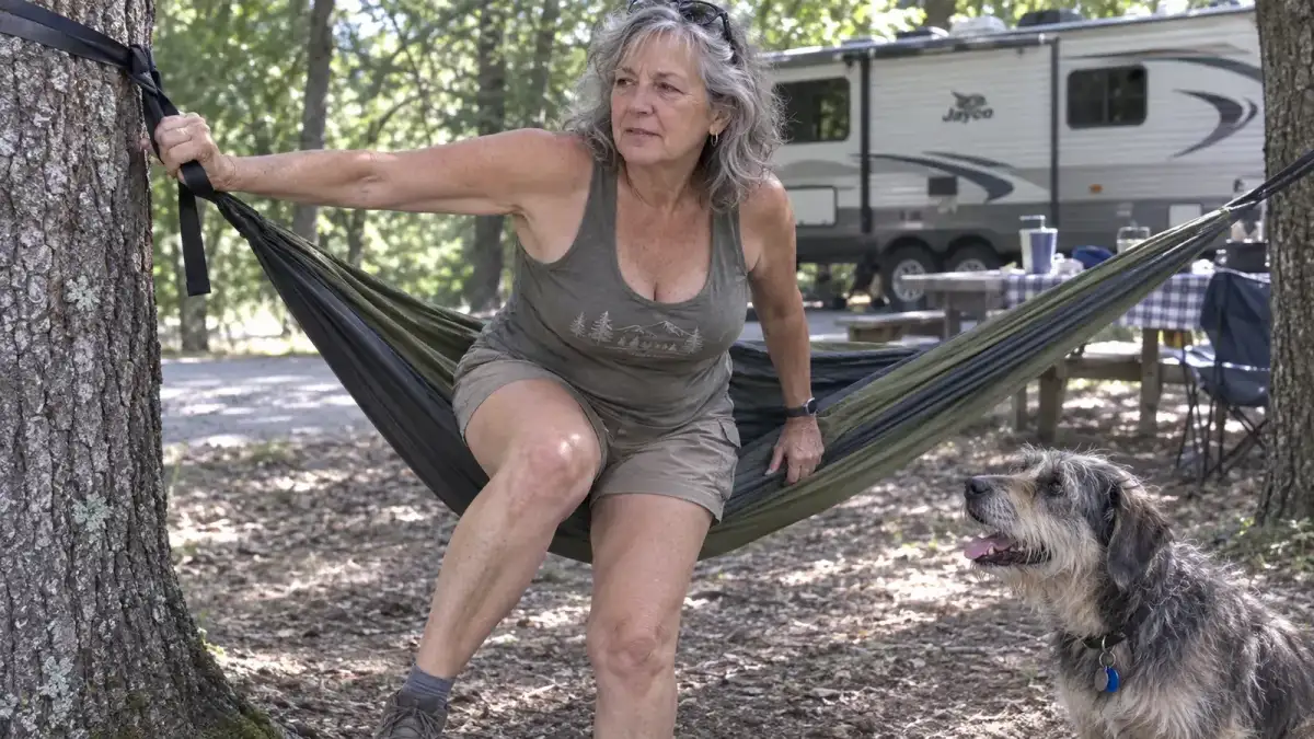 A retired woman carefully swinging her legs down to climb out of a camping hammock strung between two trees, one hand braced on the tree strap